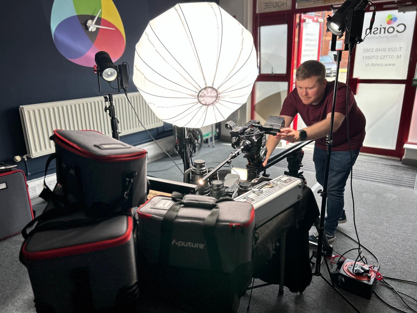 A man setting up camera equipment and lighting in a photography or video studio. The setup includes two large softbox lights, a camera mounted on a rig, and various camera accessories. The studio has dark walls with a color wheel mounted on the wall 
