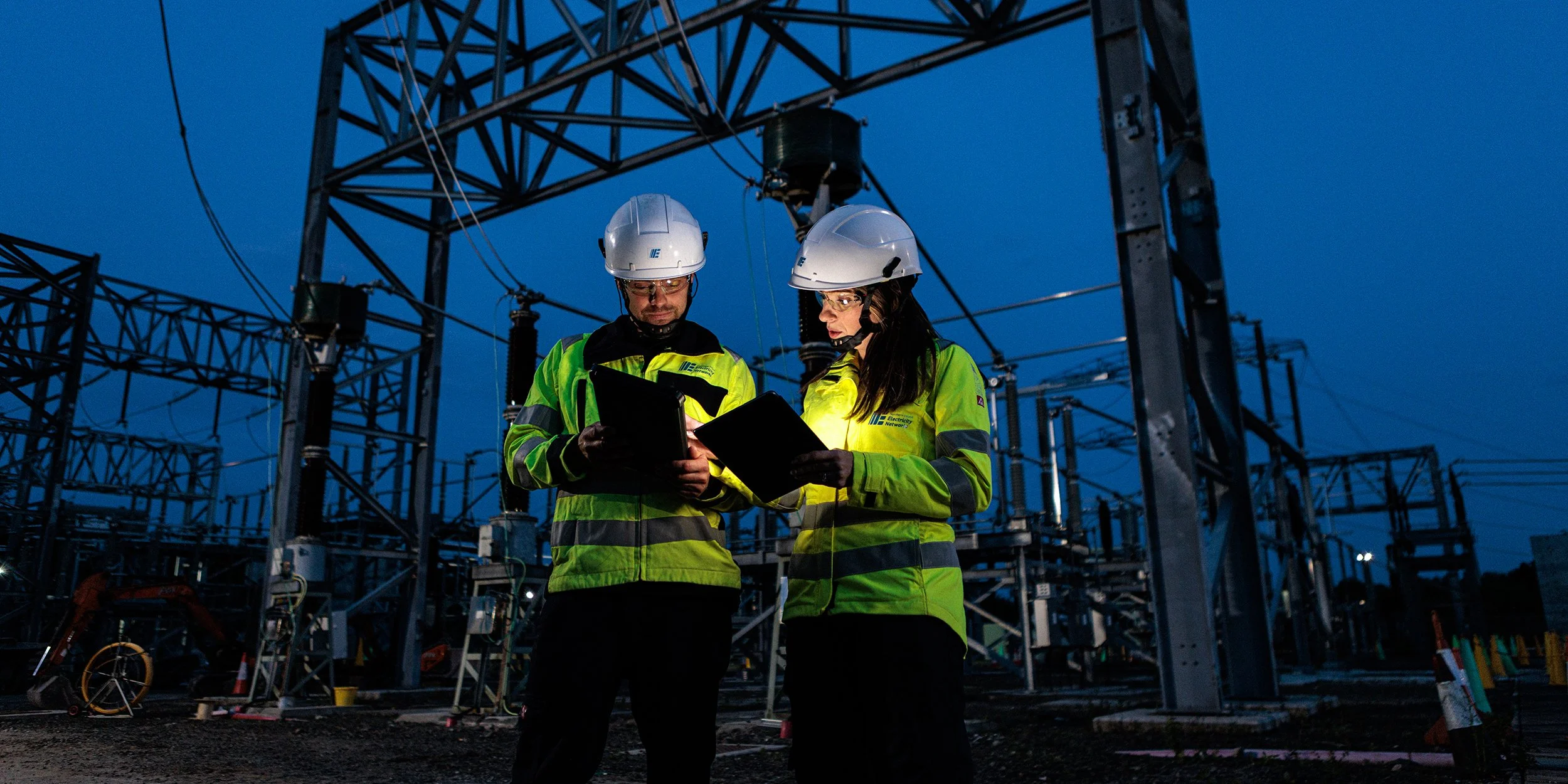 Two electrical workers wearing helmets and high-visibility jackets reviewing documents at an electrical substation during dusk.