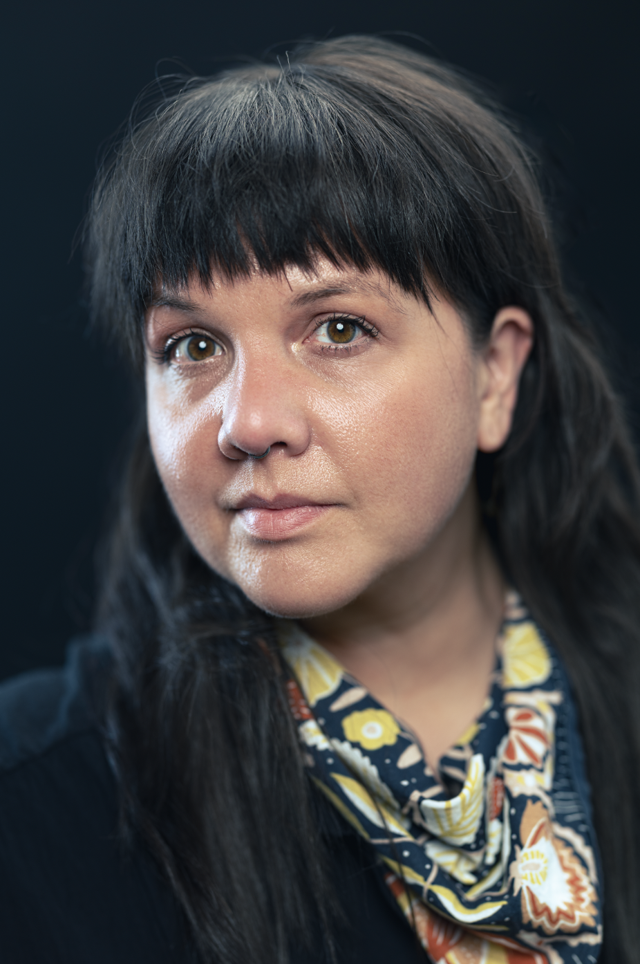 A woman with long dark hair and straight bangs is shown in a close-up portrait, looking slightly past the camera with a calm, attentive expression. She is lit softly against a dark background and wears a patterned scarf and a dark top.