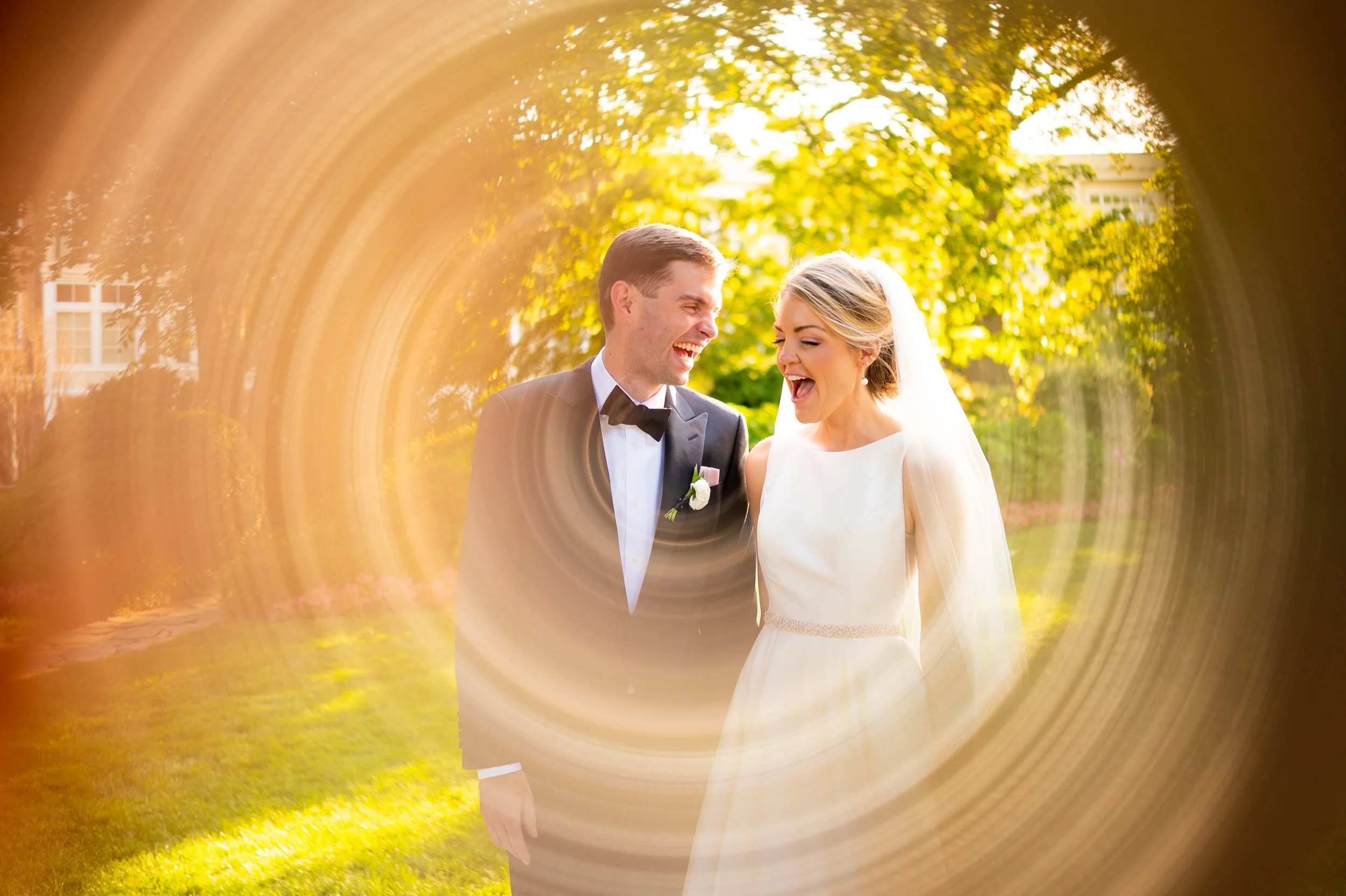 A newlywed couple smiling and laughing outdoors in a park, framed by a circular blur effect, with trees and sunlight in the background.
