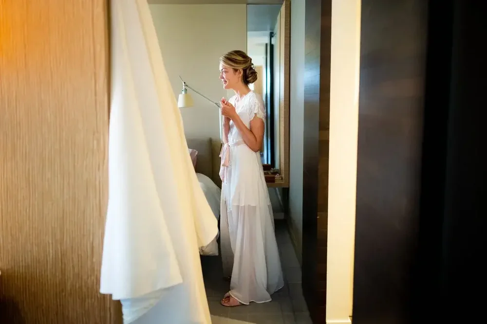 Bride in white robe standing by the window with her wedding dress hanging nearby at Meridian House