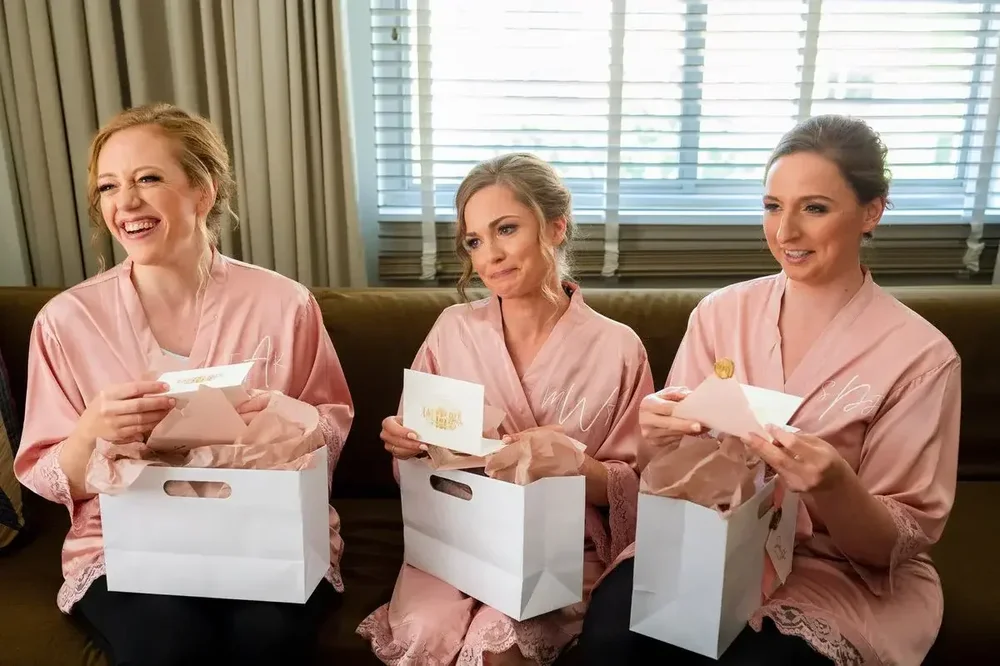 Three bridesmaids in pink satin robes opening wedding day gift bags before the Meridian House ceremony