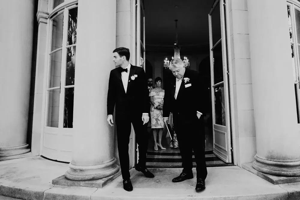 Groom and father of the bride stand at the grand columned entrance of Meridian House