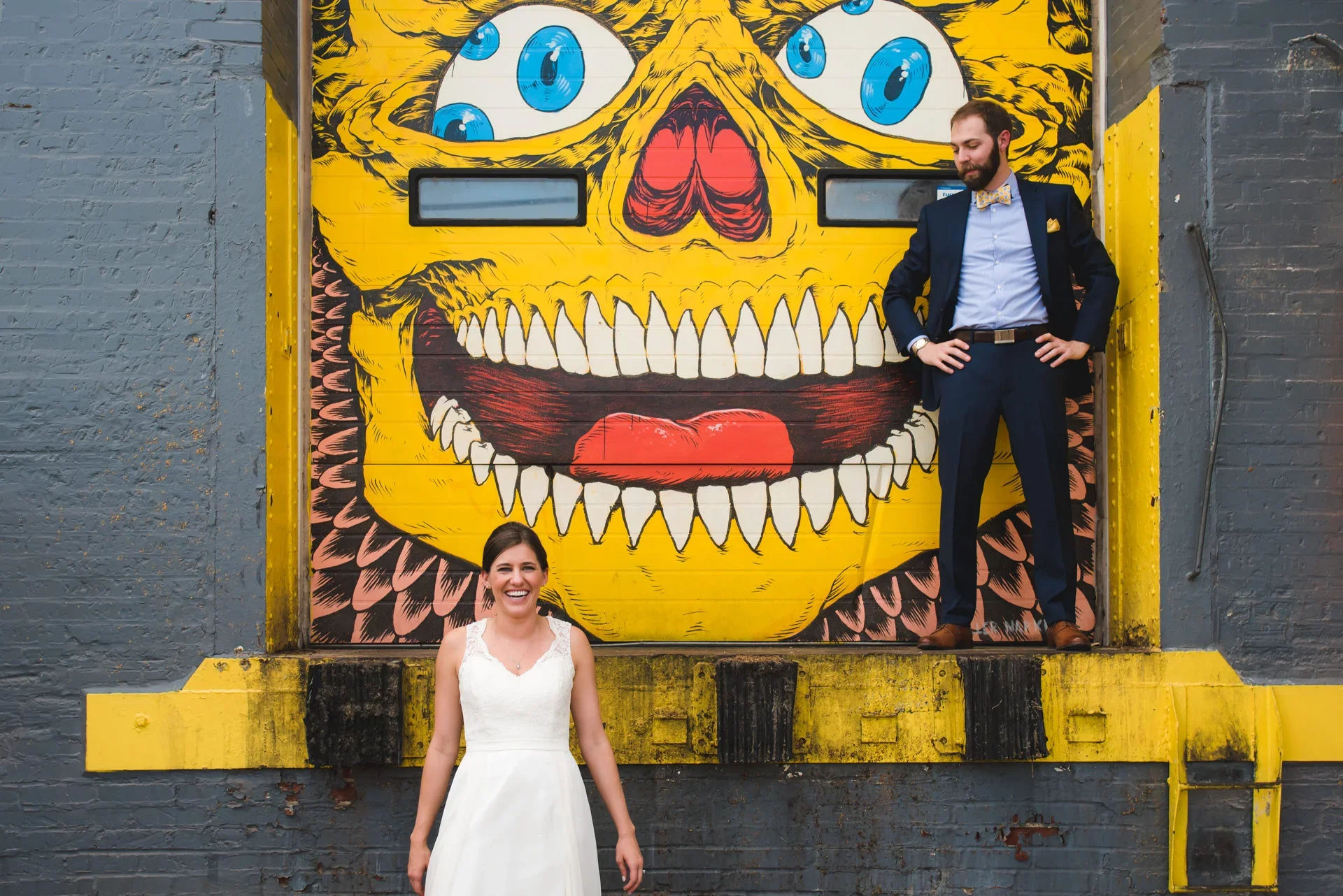 A woman in a white dress is smiling and standing in front of a gray brick wall. Behind her, a man in a suit with a bow tie and yellow pocket square is standing on a ledge in front of a large mural of a yellow monster face with blue eyes, red nose, and red tongue, featuring sharp teeth.