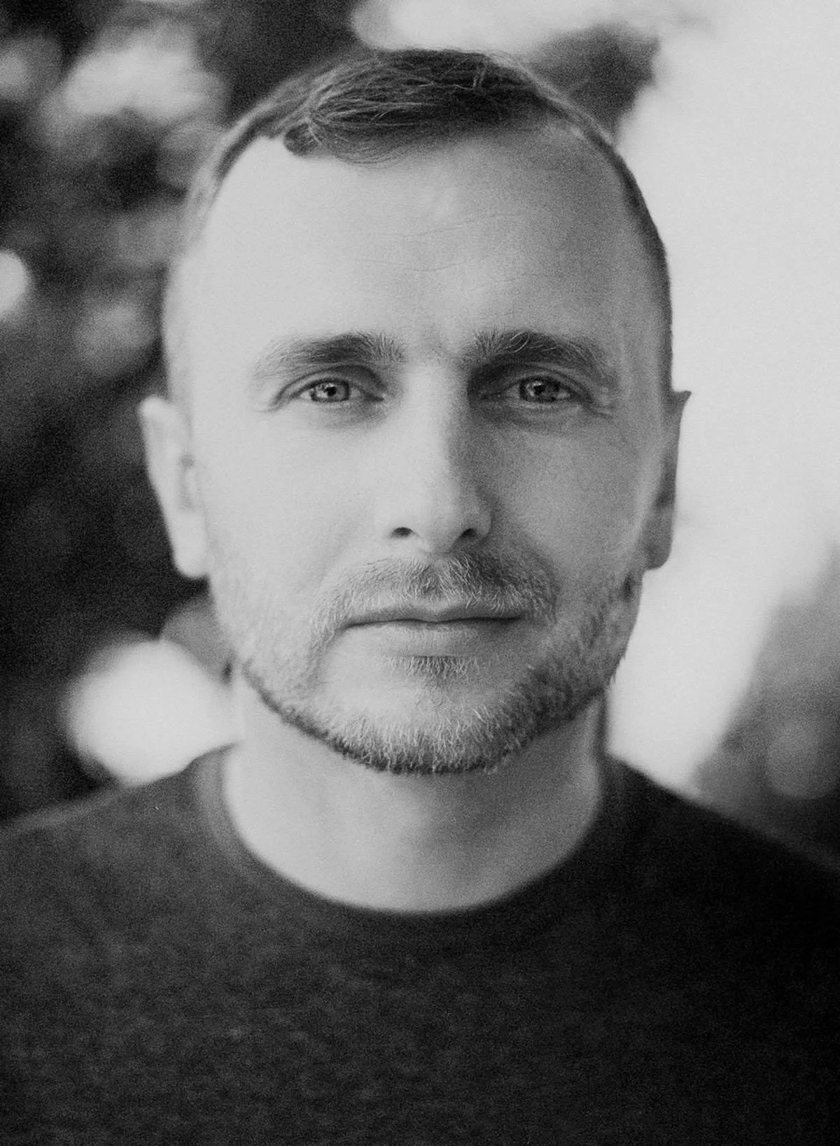 Black and white close-up portrait of a man with short hair, a beard, and light eyes, wearing a dark shirt.