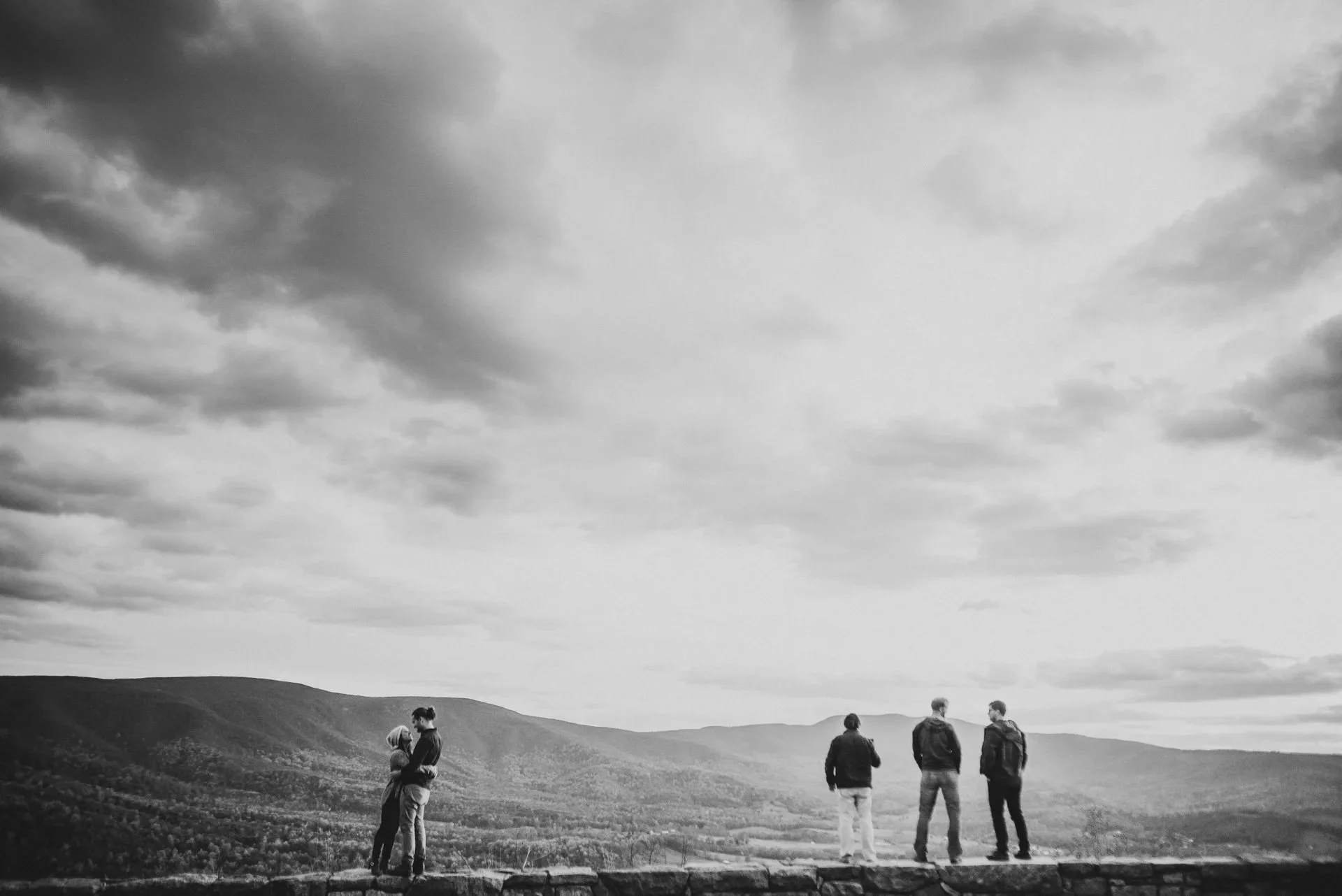 Wide scenic view of couple embracing on mountain overlook with friends nearby during Shenandoah National Park engagement session, dramatic sky over valley landscape.