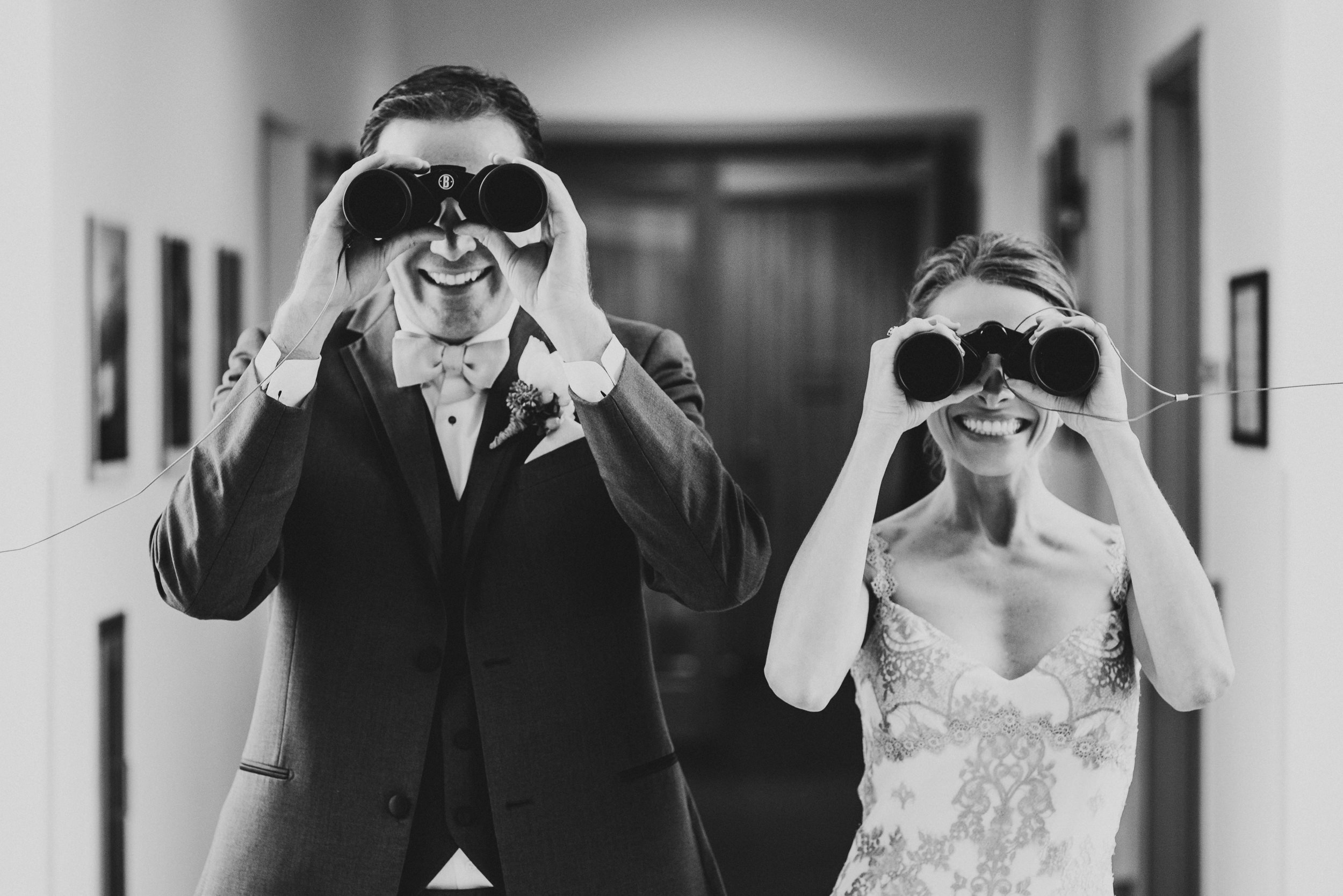 A smiling groom and bride in wedding attire holding binoculars and looking through them.