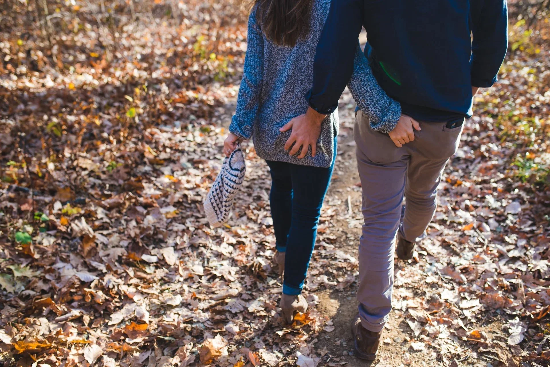 Close-up engagement photo of a couple walking hand in hand on a fall forest trail in Shenandoah National Park with autumn leaves on the ground.
