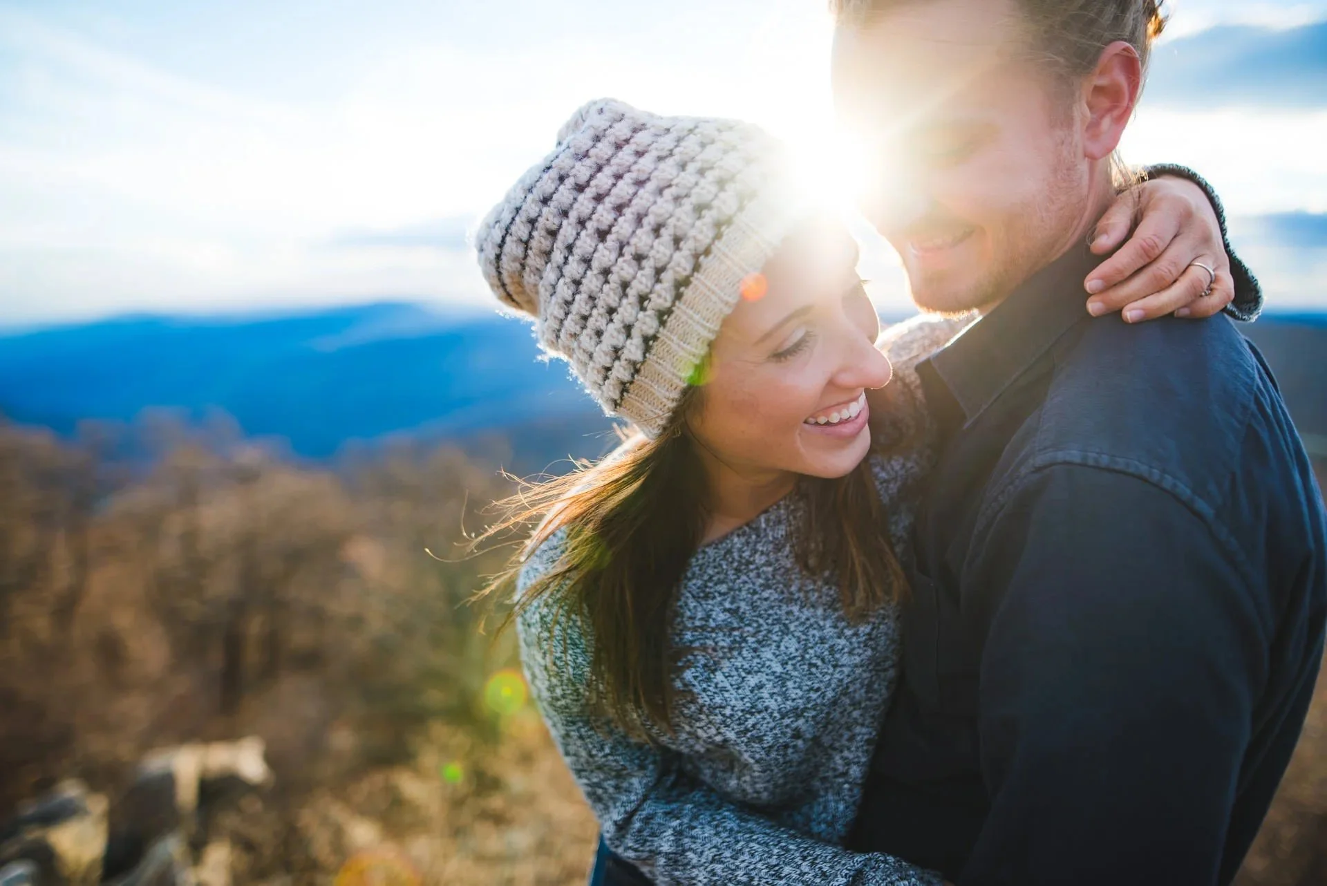 Close-up engagement photo of a couple embracing with sun flare at a mountain overlook in Shenandoah National Park with Blue Ridge Mountains in the background.