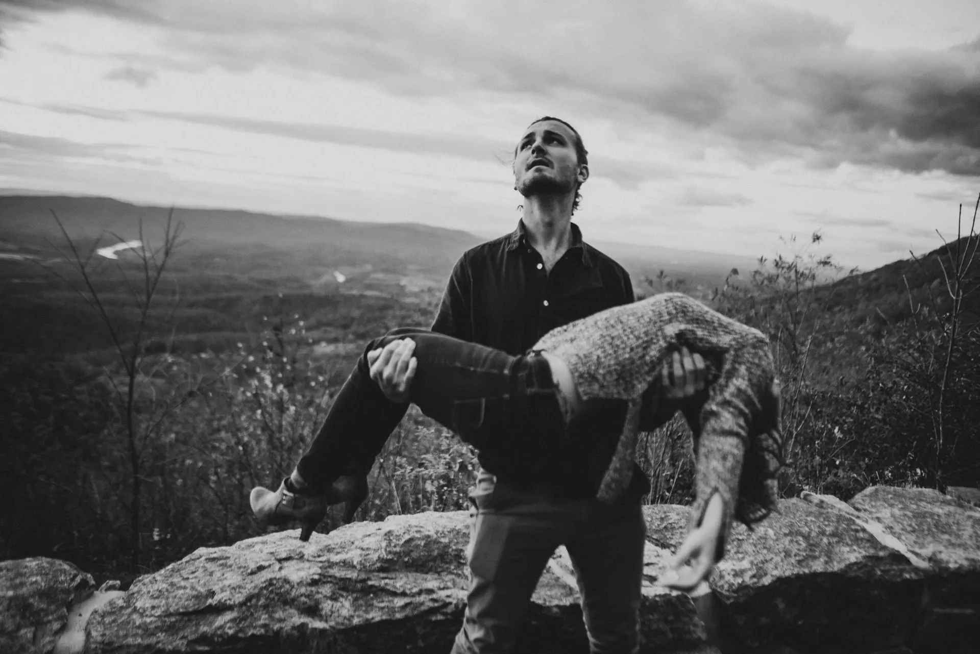 Man holding partner in his arms on a mountain overlook during Shenandoah National Park engagement session, dramatic black and white portrait with scenic valley view.