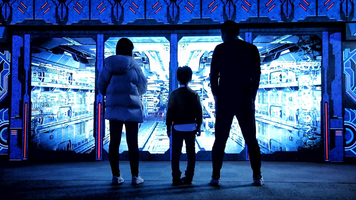 A family of three, a woman, a young boy, and a man, stands in front of a large futuristic, glowing blue digital display or virtual reality setup, viewing an immersive sci-fi scene.