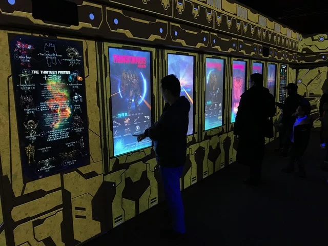 People using interactive digital exhibits with vibrant game or entertainment graphics on a yellow and black themed wall display.