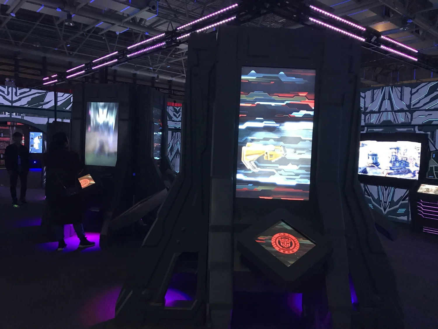 Dark room with illuminated video game screens and futuristic decor featuring purple and blue lighting, attendees playing games.