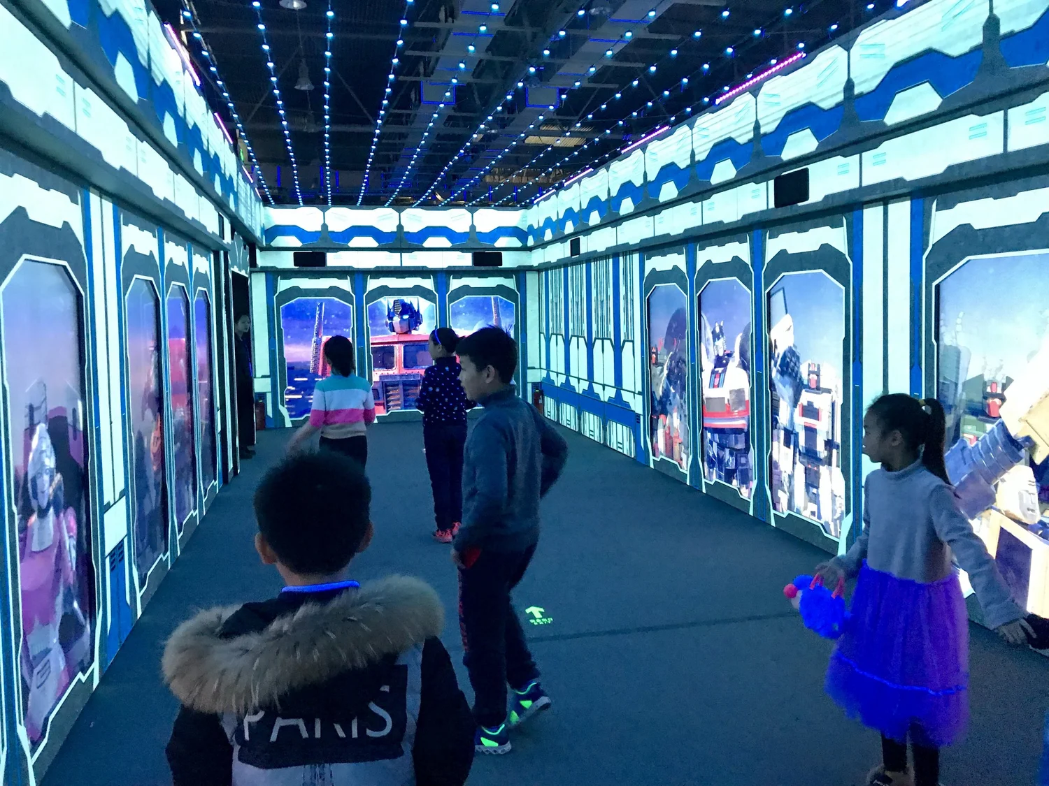 Children playing in an immersive, LED-lit virtual reality room with space-themed decor and screens displaying robots and futuristic scenes.