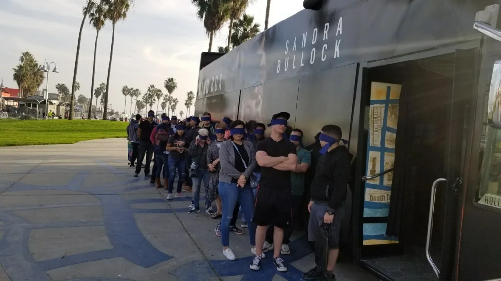 People standing and waiting in line outside a bus with the words "Sandra Bullock" and promotional posters on the side, with palm trees and a park in the background.