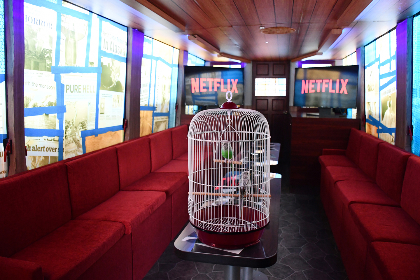 A cozy room with red upholstered seats along the walls. In the center, there is a birdcage with a small bird. The walls display newspaper-style posters, and two large screens in the back show the Netflix logo.
