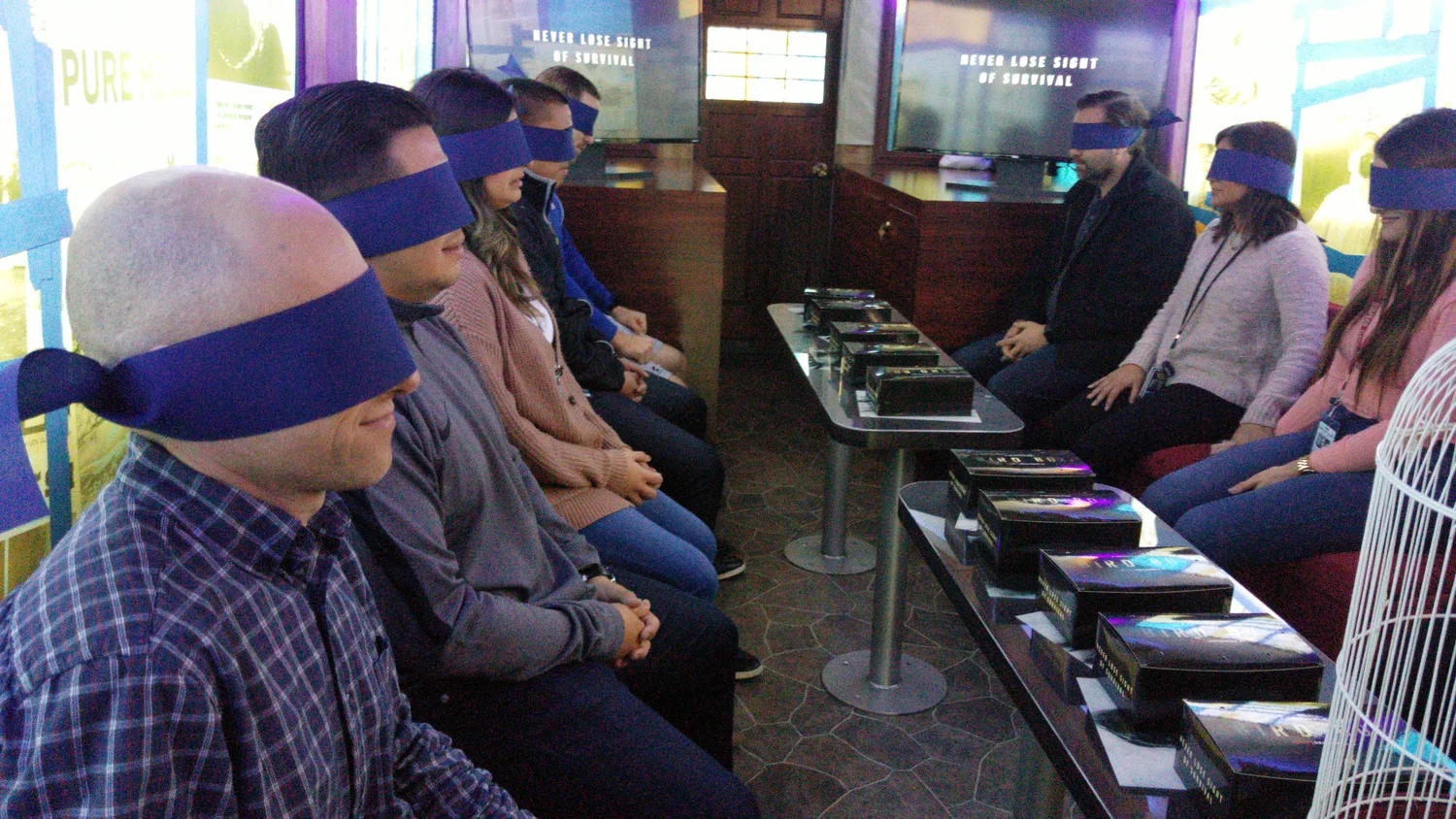 Group of people sitting in two rows with blindfolds on, engaging in an activity in a room with posters and tablets on tables.