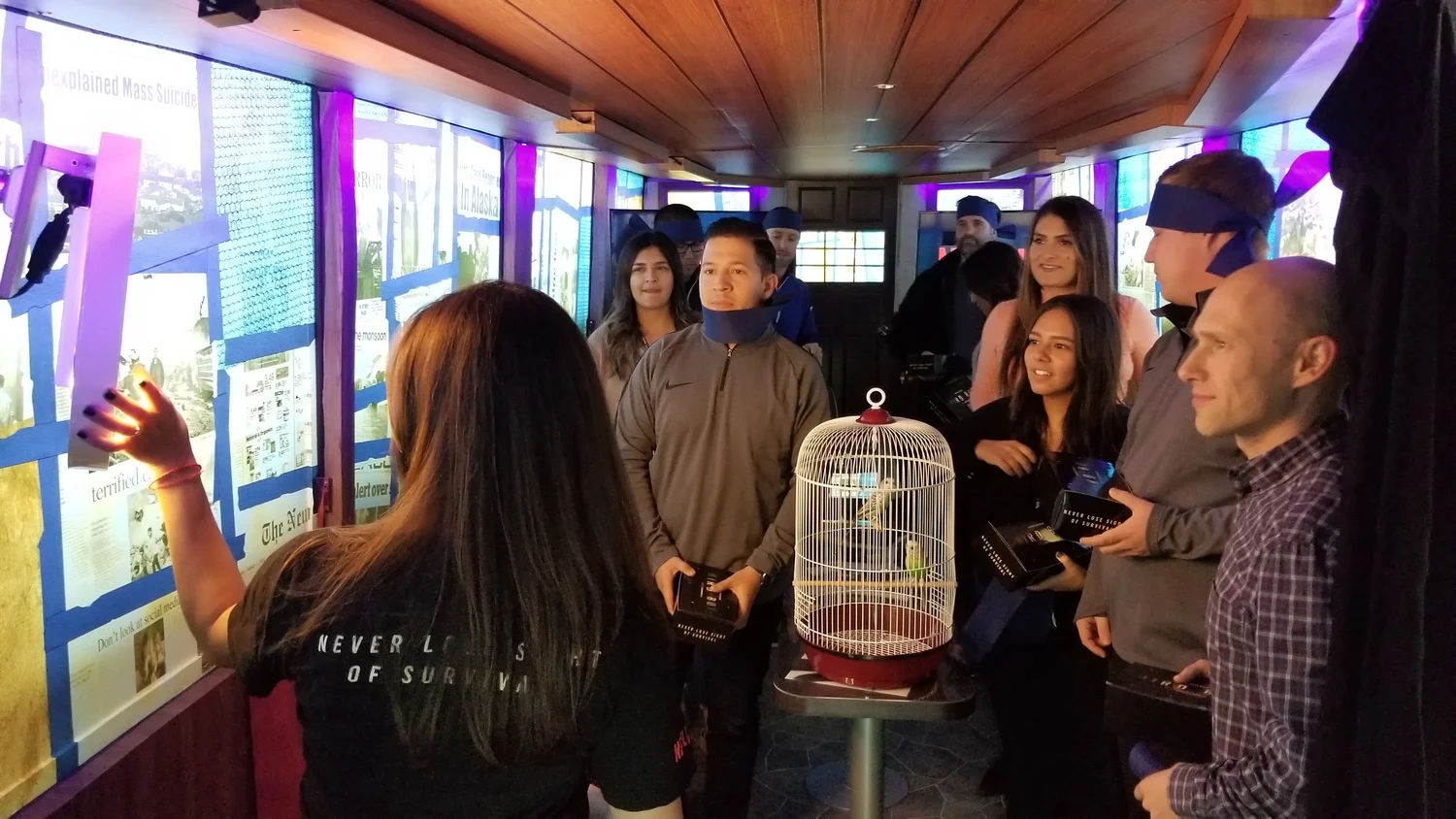 Group of people standing inside a dimly lit room, looking at display boards, with one person pointing at them. There is a birdcage with four small birds on a table in the foreground.