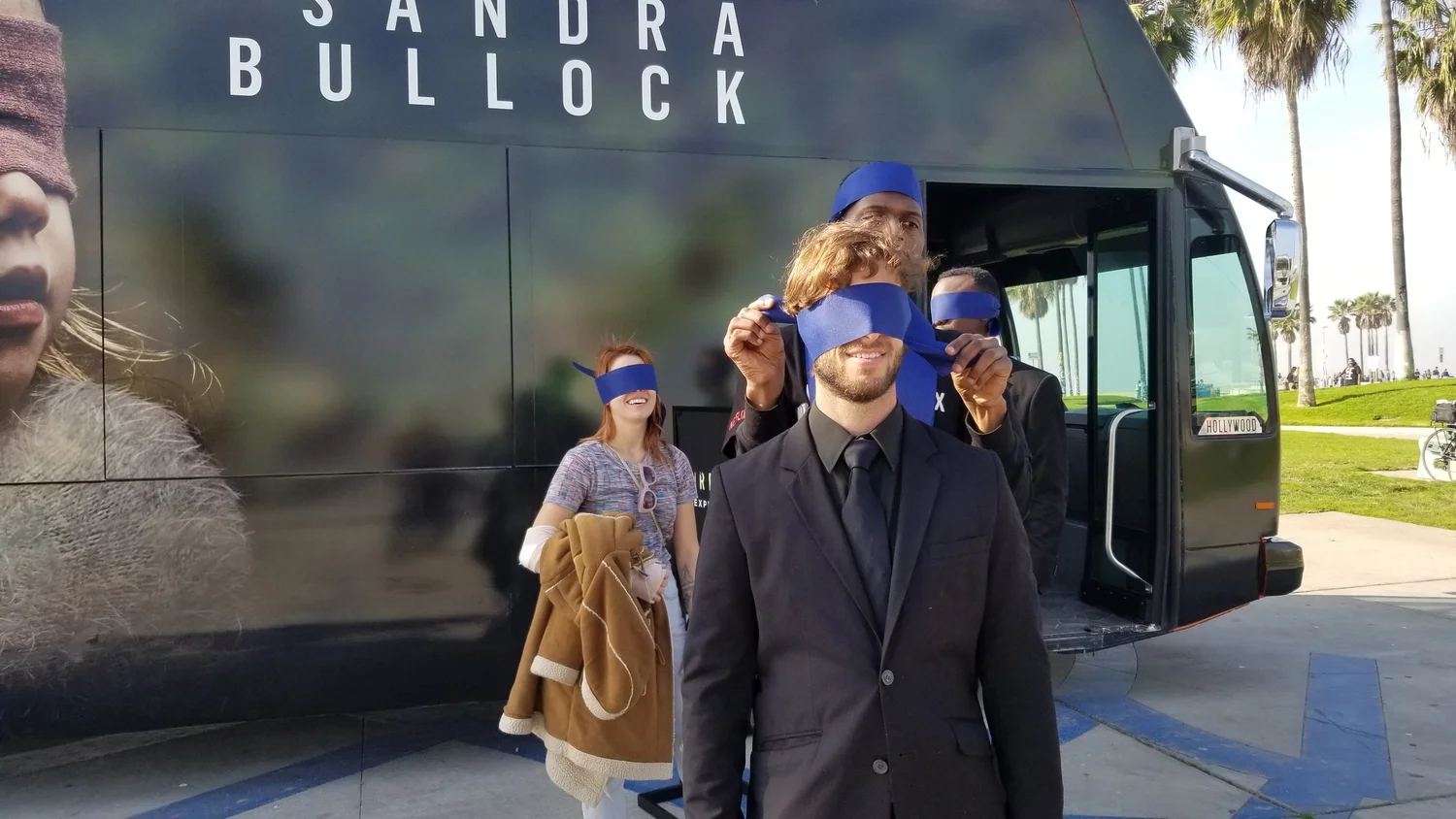 People standing outside next to a black bus with a large advertisement, with some of them blindfolded using blue cloths during daytime.