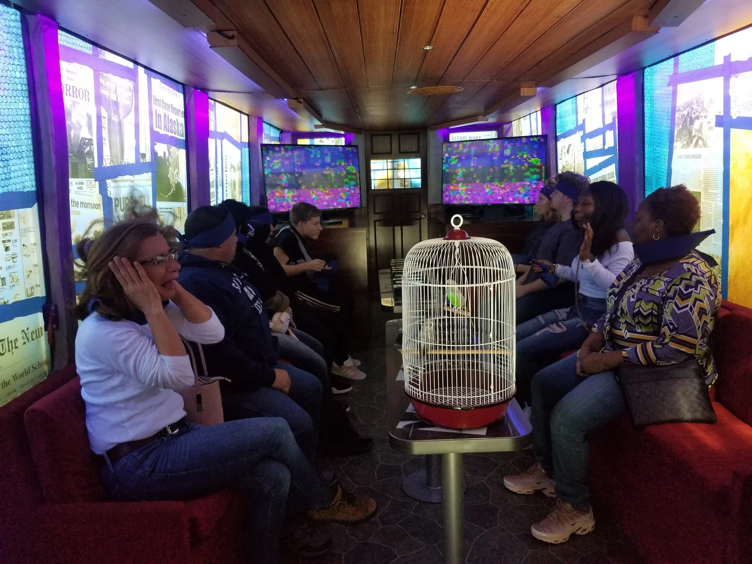 A group of people from diverse backgrounds sitting in a dimly lit room, some with headbands, watching a bird in a cage placed on a small table in front of them. The room has colorful screens and posters on the walls.