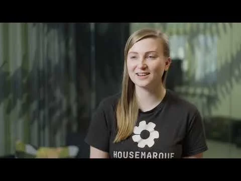 Young woman with long blonde hair, wearing a black t-shirt with a white flower and the text 'Housemarque,' sitting in a modern indoor setting with greenery in the background.