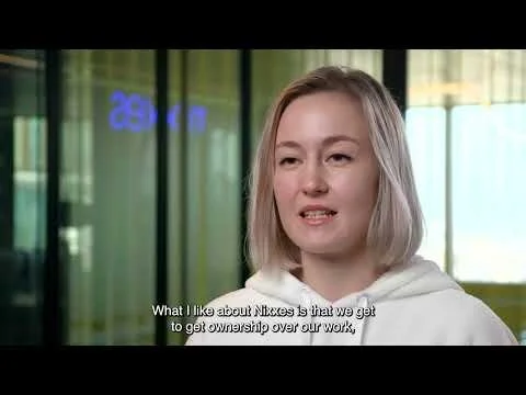 A young woman with blonde hair speaking in an interview setting with a blurred modern office background. Subtitles read: 'What I like about Nixes is that we get to get ownership over our work.'