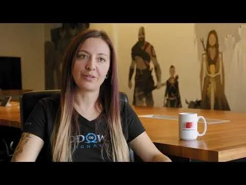 A woman with long hair sitting at a wooden table with a white mug, in an office or studio setting with artwork on the wall behind her.