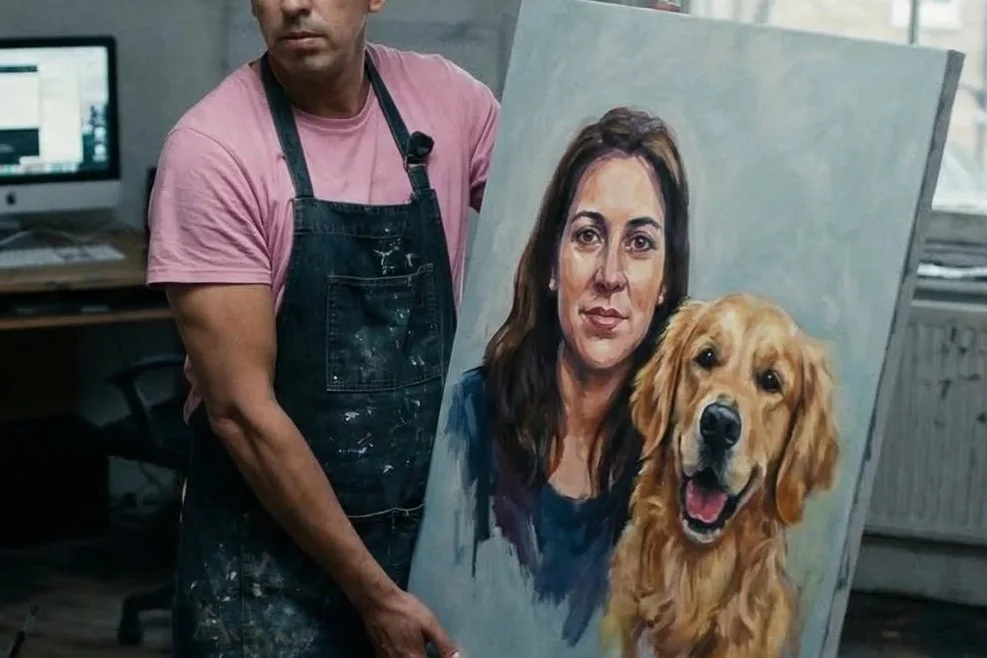 An artist stands in an art studio with a painting of a woman and a golden retriever dog. The artist wears a pink t-shirt and a black apron with paint splatters. The painting depicts a woman with brown hair and a golden retriever barking happily.