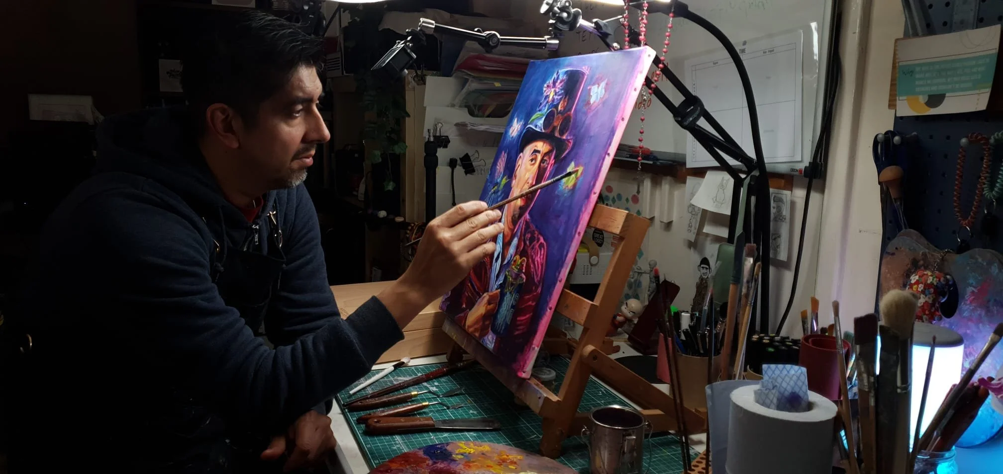 Man painting a colorful portrait on a canvas held on an easel in a dimly lit art studio surrounded by art supplies and decorations.