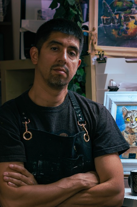A man with short black hair and a goatee, wearing a black t-shirt and apron, standing with arms crossed in an art studio surrounded by paintings and art supplies.