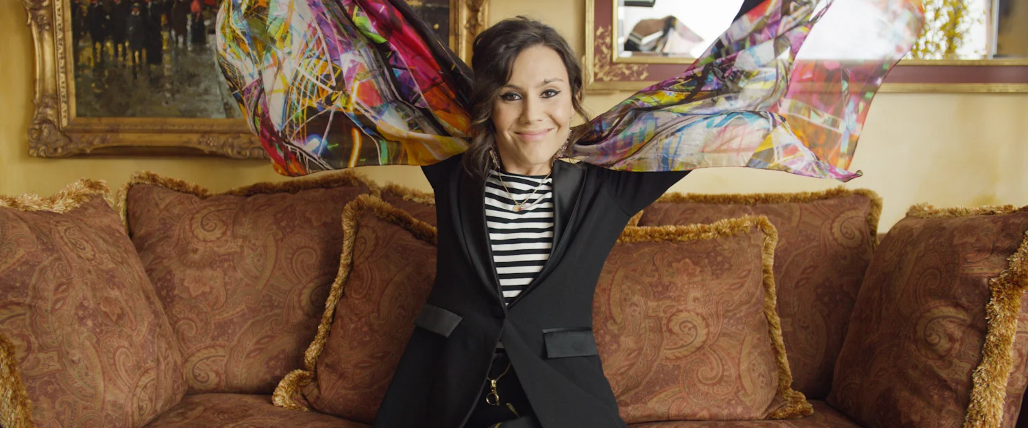 A woman smiling and sitting on a sofa, holding colorful fabric in the air behind her. She is wearing a black blazer over a black and white striped shirt, in a room with paintings and a window in the background.