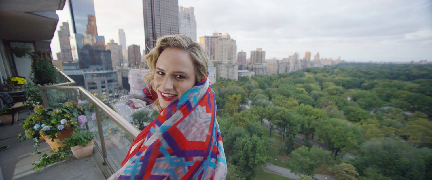 Woman with blonde curly hair and red lipstick smiling on a balcony with city skyline and park in the background, wearing a colorful geometric shawl.