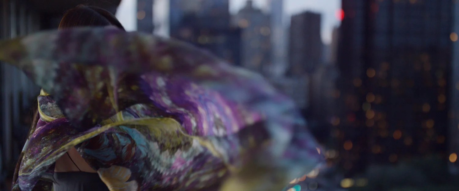 A person partially obscured by a colorful, flowing scarf stands in an urban setting with city lights and skyscrapers in the background.