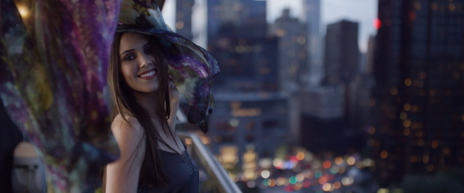 A woman with long dark hair, wearing a dark dress and a colorful umbrella, smiling at the camera with a city skyline and blurred city lights in the background at dusk.