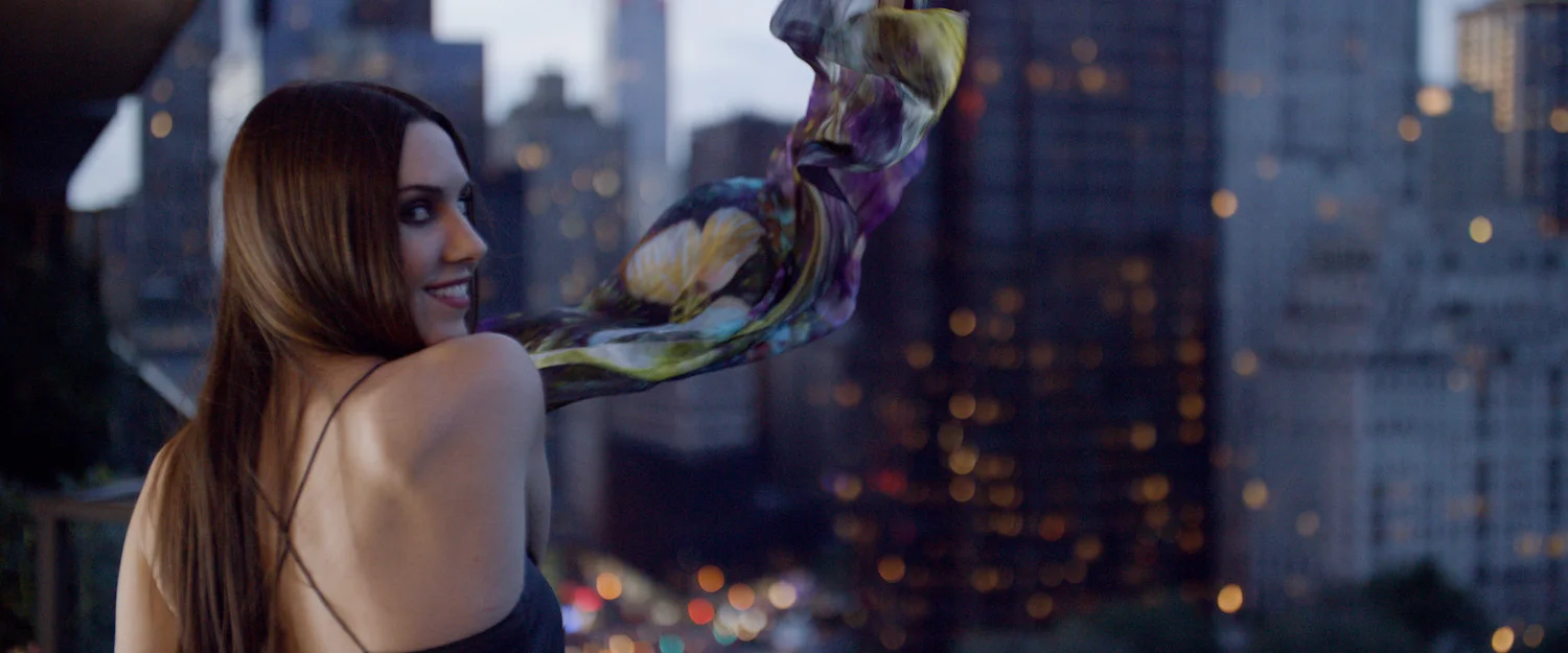 A woman with long brown hair in a black dress with spaghetti straps, smiling and looking over her shoulder, with a colorful scarf flowing in the wind, against a cityscape background during dusk.