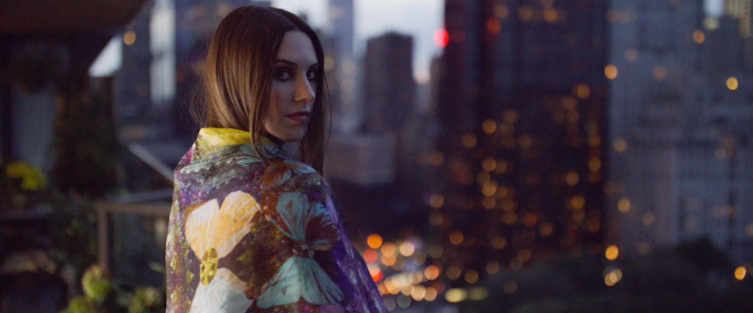 Young woman with brown hair and makeup looking over her shoulder, standing outdoors at dusk, wearing a colorful floral jacket with city skyscrapers in the background.