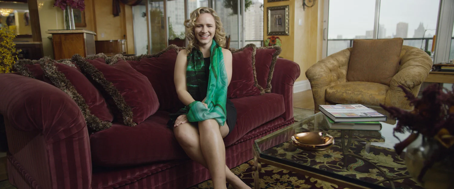 A woman with curly blonde hair sitting on a plush red velvet sofa, smiling and wearing a black dress with a green scarf, in a well-decorated living room with large windows and a view of a city skyline.