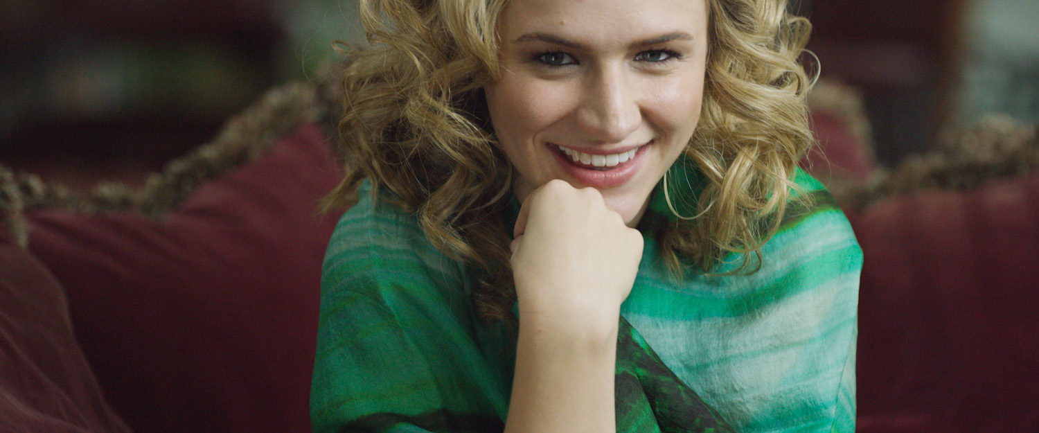 Close-up of a woman with curly blonde hair and a green blouse, smiling with her chin resting on her hand, sitting on a maroon couch.