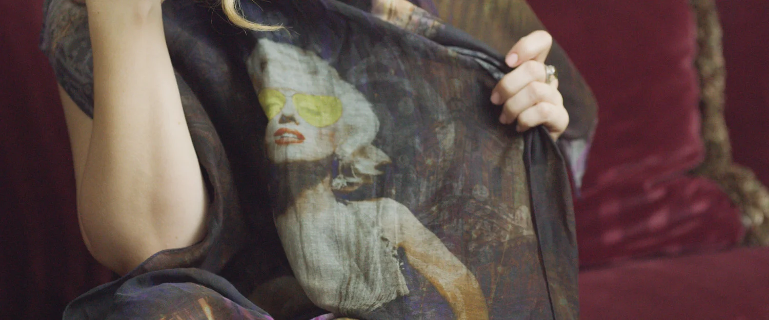 Person wearing a sheer black shirt featuring a vintage-inspired portrait of a woman with sunglasses, red lips, and curly hair, sitting on a red velvet couch.