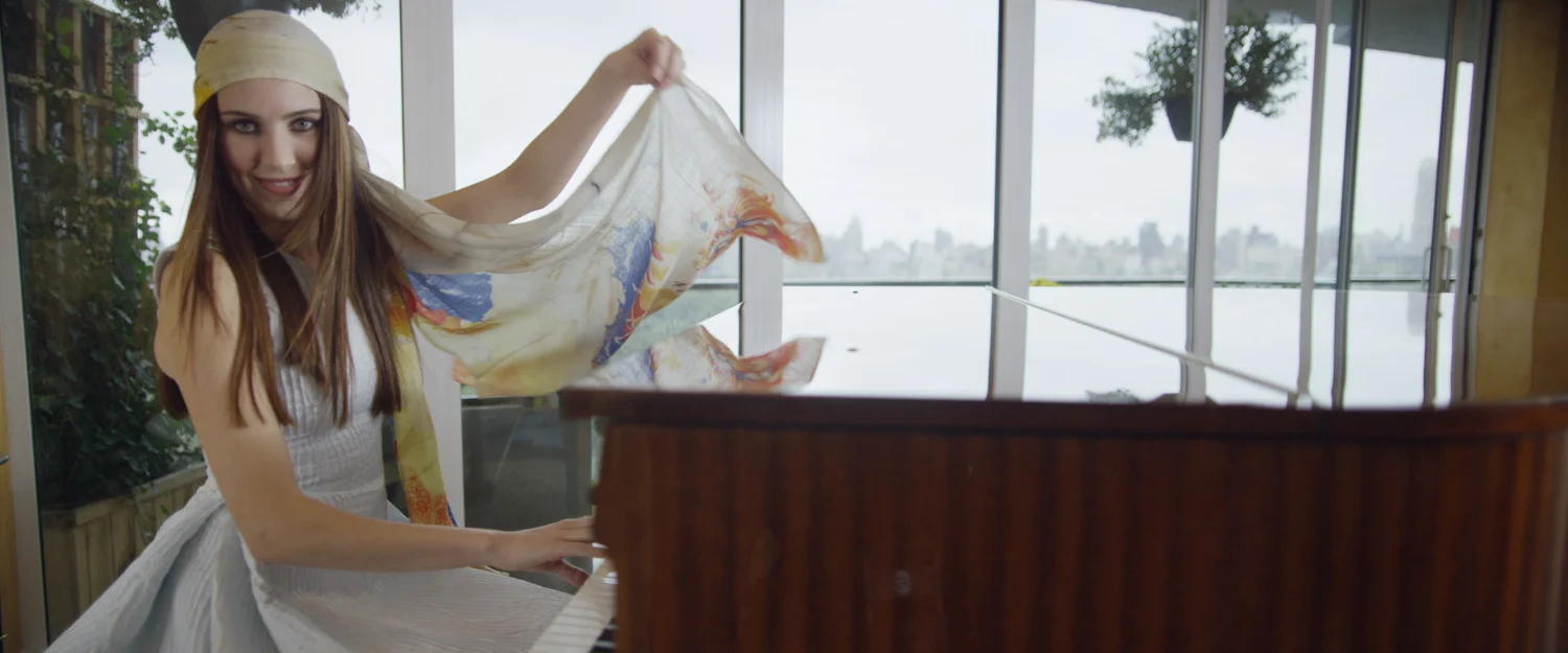 Woman with long brown hair, wearing a yellow headscarf and white dress, smiling while playing the piano in a bright room with large windows and cityscape view.