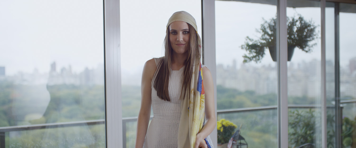 A woman with long brown hair wearing a cream-colored headscarf and white sleeveless dress standing indoors near large glass windows with a cityscape and park view in the background.