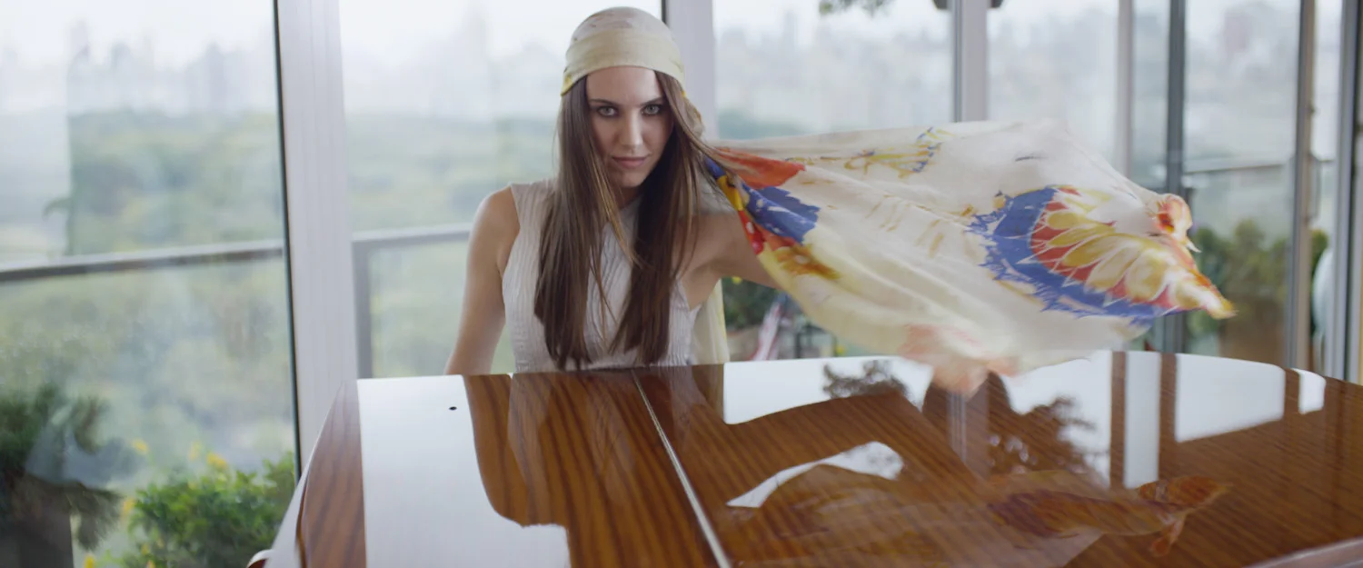 A woman with long brown hair wearing a headscarf, sitting at a polished wooden table in a bright room with large windows, with a scarf flowing behind her.