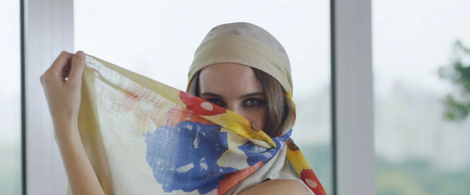 A woman with brown hair wearing a headscarf and holding a colorful scarf in front of her face, with only her eyes visible.