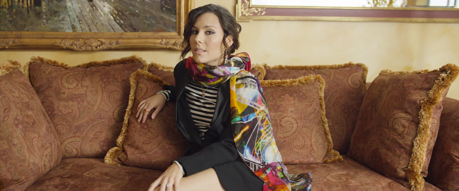A woman sitting on an ornate peach-colored couch with gold accents, wearing a black blazer, striped shirt, and a colorful scarf around her neck, in a room with framed artwork on the wall.