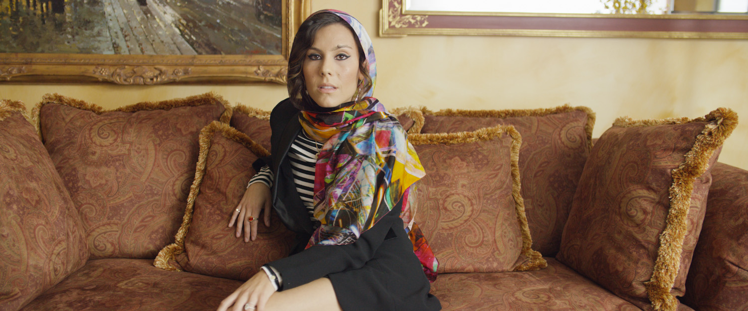 A woman sitting on a patterned sofa with multiple pillows, wearing a colorful headscarf and a black dress with striped sleeves, looking at the camera in a well-decorated room.