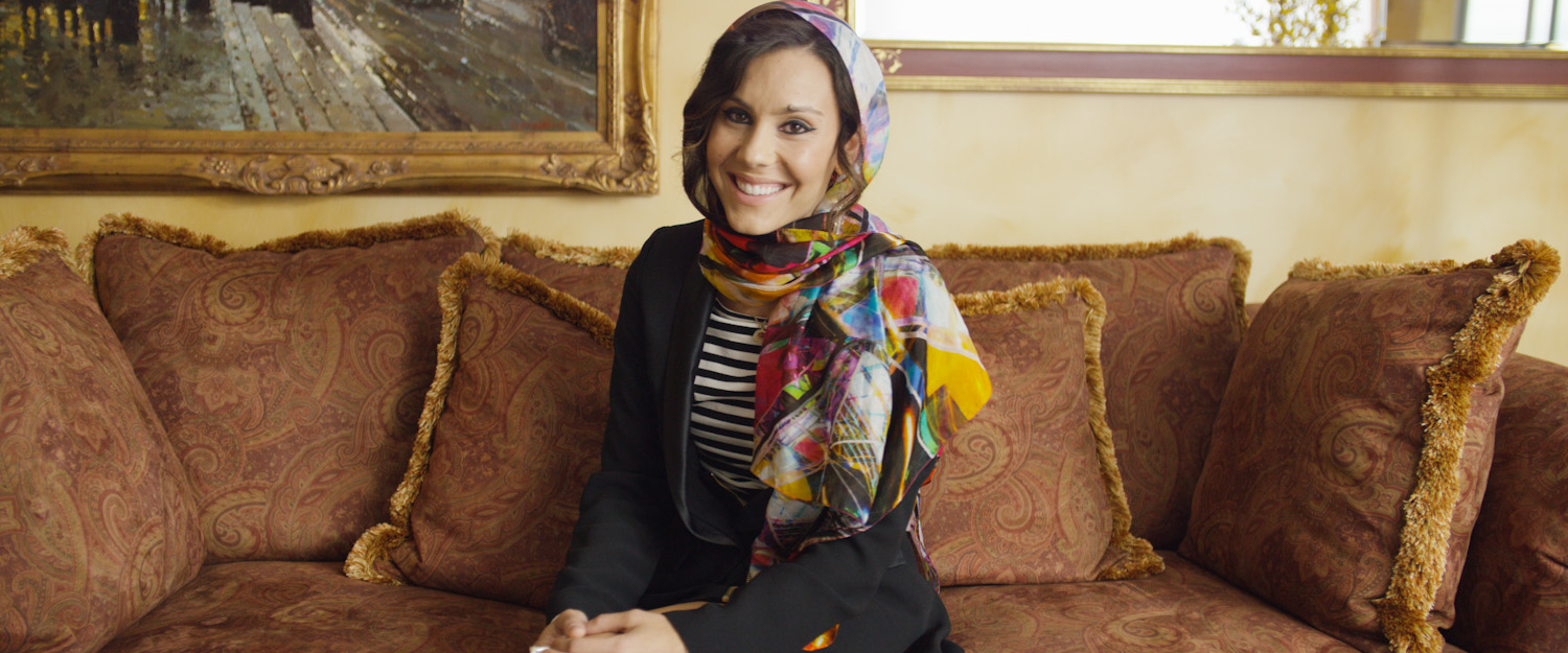 A woman with dark hair smiling, wearing a colorful headscarf, a black blazer, and a striped shirt, sitting on a patterned brown sofa in a warmly lit room with artwork on the wall behind her.