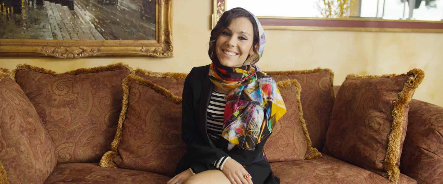 A woman sitting on a patterned brown couch with decorative pillows, smiling at the camera, wearing a colorful scarf, a striped shirt, and a black blazer in a cozy indoor setting.