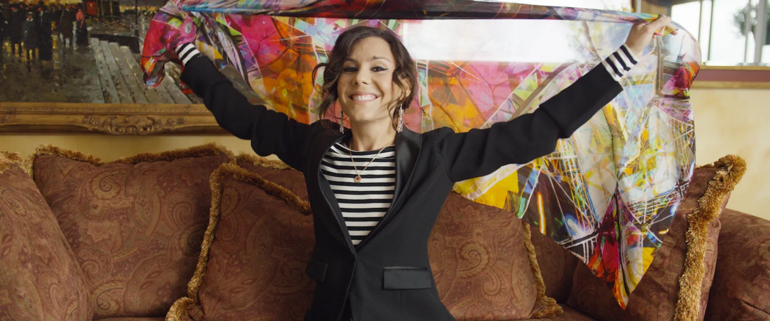 A woman with curly hair smiling and holding colorful fabric or scarf behind her head, sitting on a brown sofa in a room with artwork on the wall.