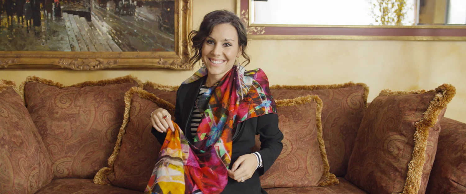 A woman with dark brown curly hair smiling and sitting on a brown sofa, holding a colorful scarf. Behind her are paintings and a mirror on the wall.