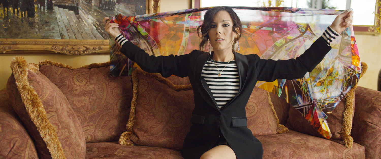 A woman sitting on a patterned couch, holding a colorful, semi-transparent scarf with both hands stretched out. She is wearing a black blazer, striped shirt, and jewelry, with dark hair styled in loose waves.